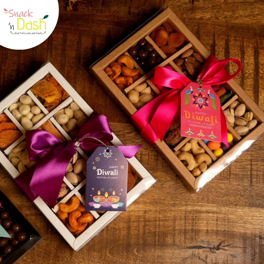 Two Diwali-themed gift boxes filled with assorted dried fruits, nuts, and chocolates, tied with bright pink and purple ribbons and decorated with festive tags, displayed on a wooden table. 