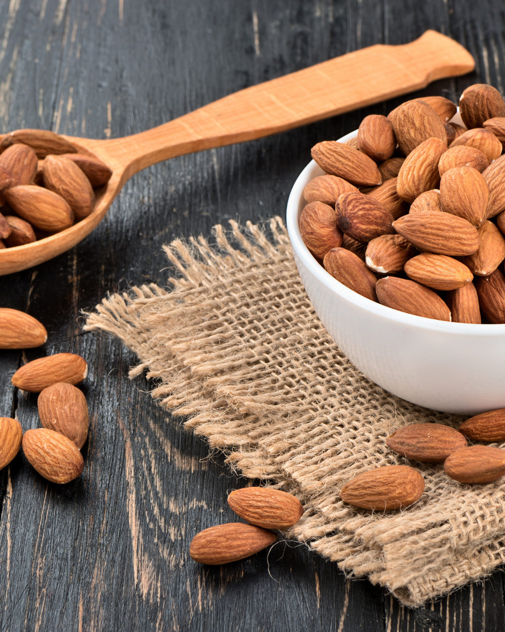 Almonds in a white bowl and on a wooden spoon with a burlap cloth on a dark surface, suitable for baking 