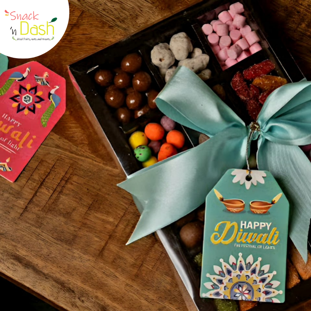 A Diwali-themed chocolate gift box filled with assorted chocolates, pink marshmallows, dried fruits, and colorful candies, tied with a large mint green ribbon and decorated with a 'Happy Diwali' gift tag. A pink 'Happy Diwali' card is placed beside the box on a wooden table.