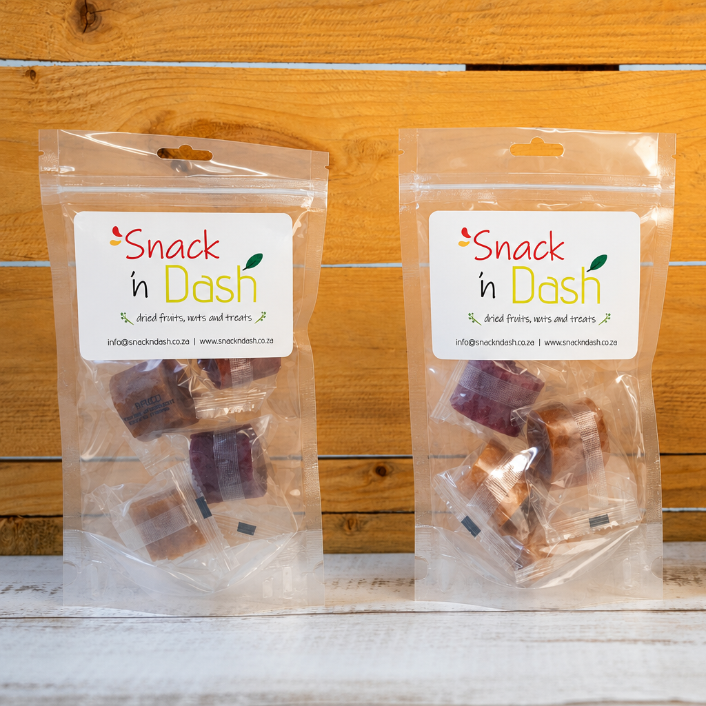 Three clear plastic bags of 'Snack in Dash' natural fruit and nut snacks arranged on a white wooden surface against a light wood plank background.