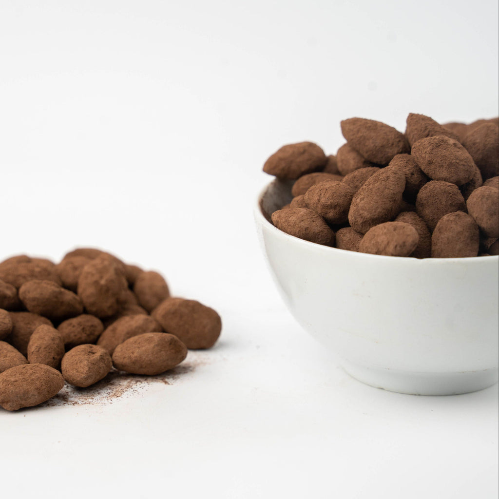 Almonds coated in cocoa powder, shown with a small white bowl on the right and a loose pile on the left against a clean white background.