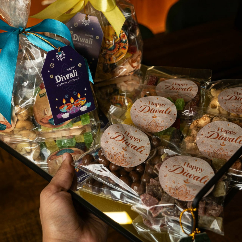 A hand holds a glass tray filled with Diwali-themed packaged treats. The arrangement includes clear plastic bags with yellow and blue ribbons, round tags reading 'Happy Diwali,' and assorted sweets and snacks visible through the wrapping.