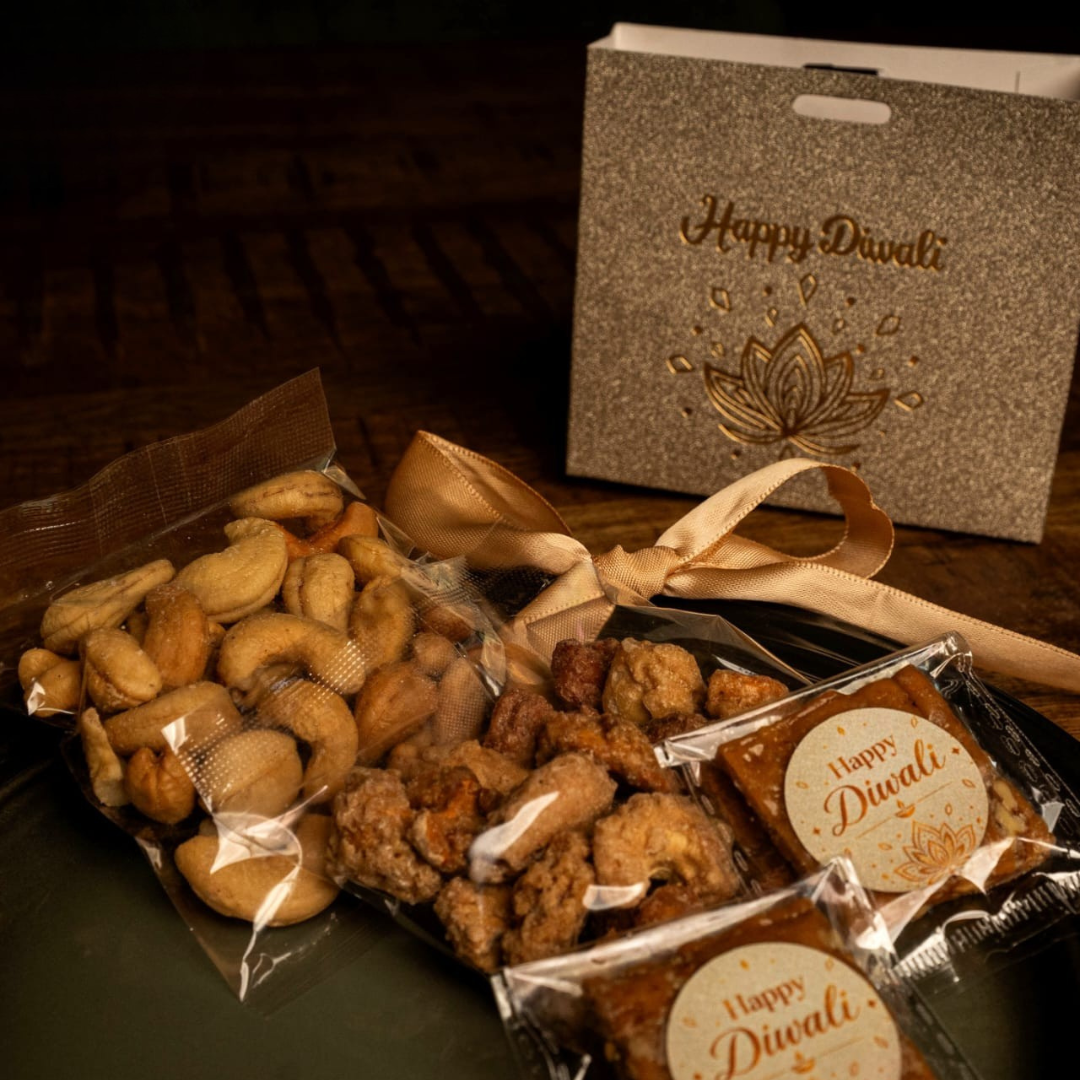 A Diwali gift set featuring assorted snacks including cashews, fried snacks, and packaged sweets, arranged on a dark tray with a glittery 'Happy Diwali' box in the background.