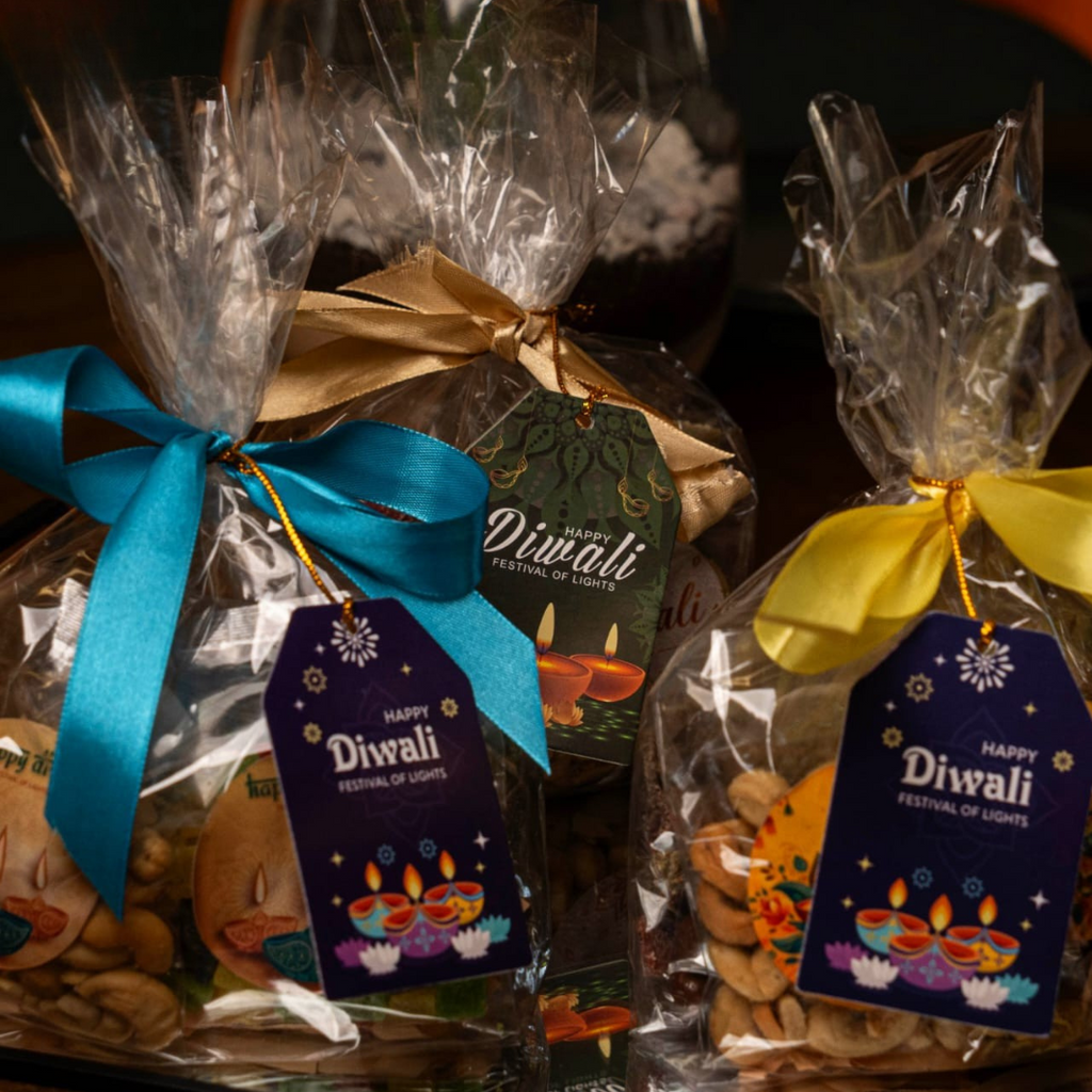 Two Diwali gift baskets filled with nuts and sweets, wrapped in clear cellophane and tied with blue and yellow ribbons. Each package has a decorative 'Happy Diwali' tag and is displayed on a dark tray.