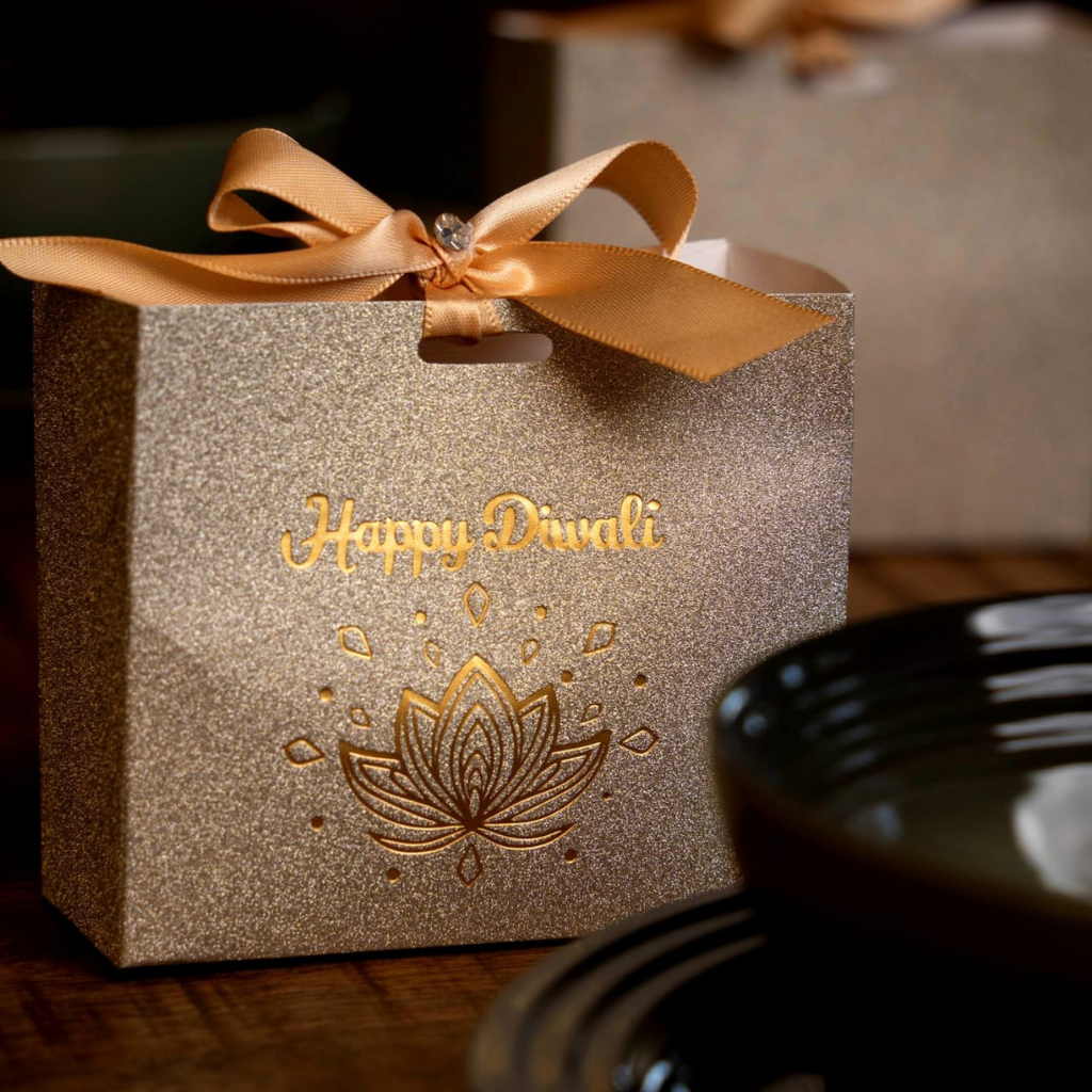 A glittery gold Diwali gift box with a gold ribbon bow and 'Happy Diwali' text, set on a wooden table with a blurred bowl in the foreground.