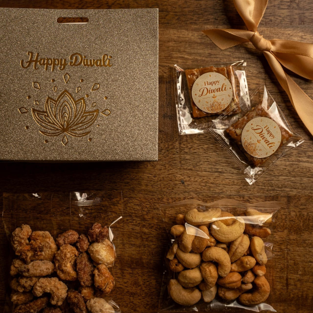 A Diwali gift set arranged on a dark wooden table, featuring a textured metallic silver box embossed with 'Happy Diwali' and a lotus design, two individually wrapped sweets with round labels, and two clear bags of mixed nuts and candied fruits. A gold satin ribbon is tied in the top right corner.