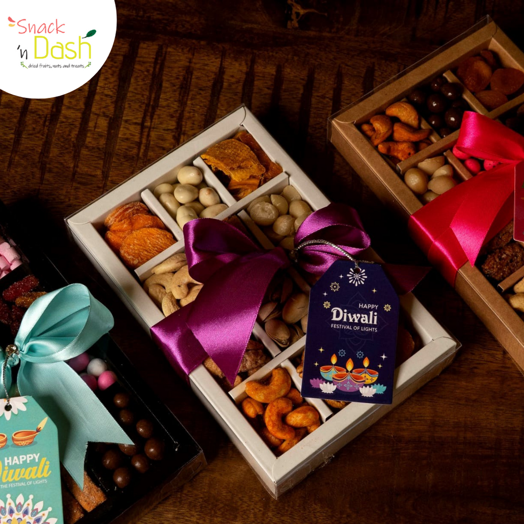 Three Diwali-themed snack gift boxes arranged on a dark wooden table. The central open box contains assorted dried fruits, nuts, and sweets tied with a purple ribbon and a 'Happy Diwali' tag.